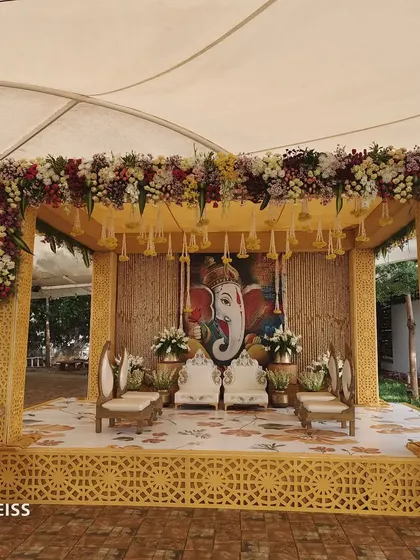 An Indowestern wedding mandap design featuring a Ganesha painting as the centerpiece. The structure uses yellow latticework and is topped with a dense arrangement of flowers, blending traditional and modern styles.