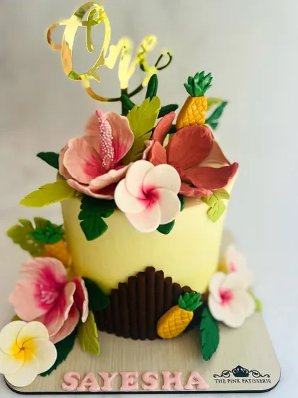 Celebrating a first birthday with a vibrant tropical-themed cake. This detailed creation features handcrafted sugar hibiscus flowers, plumeria, and miniature pineapples, all surrounding a custom gold acrylic "One" topper.