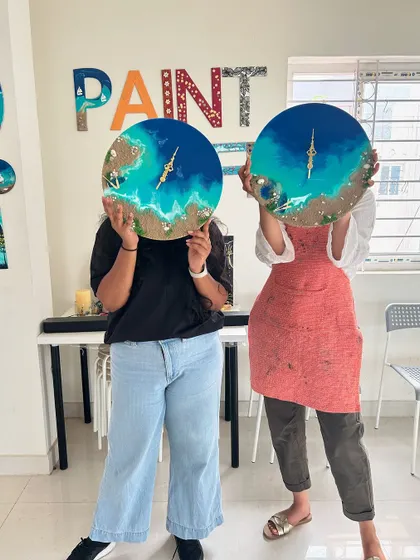 Two participants playfully hold their finished beach clocks in front of their faces, showing the fun atmosphere of our workshops.