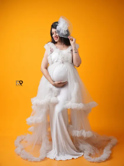 A playful and elegant pose. The mom-to-be looks radiant in a white ruffled gown, with a delicate fascinator adding a touch of vintage charm. The bright yellow background adds a burst of energy.