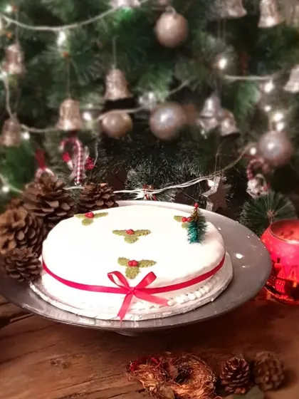 A simple, elegant Christmas cake with a smooth white icing, decorated with holly and a tiny Christmas tree. Set against the warm glow of a Christmas tree, it captures the cozy feeling of the holidays.