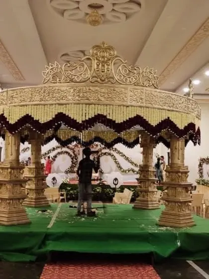 A magnificent circular wedding mandap being set up by my team. The golden structure is intricately carved, and the floral arrangement of white and maroon flowers adds a touch of elegance and grandeur.
