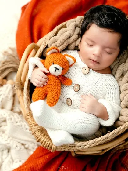 Warm, earthy tones create a cozy atmosphere for this newborn portrait. The baby, dressed in a knitted outfit, cuddles a teddy bear while nestled in a woven basket.