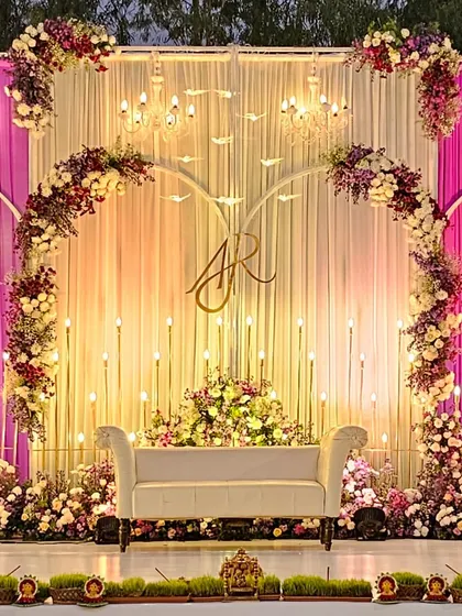 A romantic and sophisticated reception stage with a custom monogram. The design features beautiful floral arches, warm candle-like lighting, and elegant drapery in shades of pink and cream.