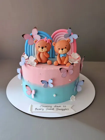 Counting down to beary sweet snuggles. This baby shower cake for twins features two teddy bears, pink and blue rainbows, and delicate butterflies.