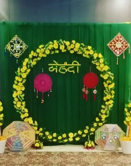 A simple Mehendi photo booth with a circular frame of yellow flowers against a green backdrop. The decor includes hanging dreamcatchers and colorful umbrellas.