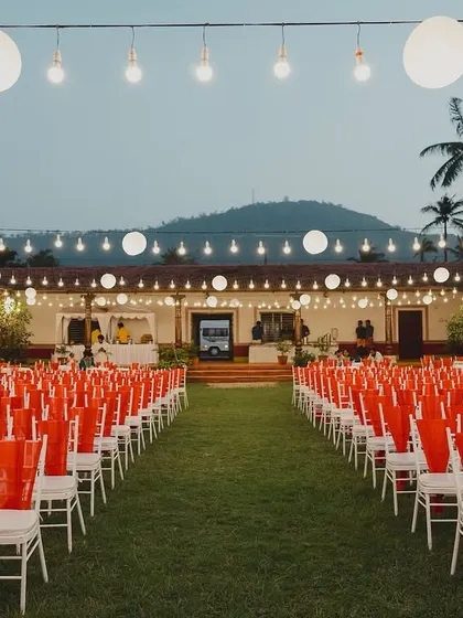 The guest seating for a Sangeet under the stars. The vibrant orange chair covers add a pop of color, while strings of paper lanterns and bulbs create a festive and magical ambiance against the twilight sky.