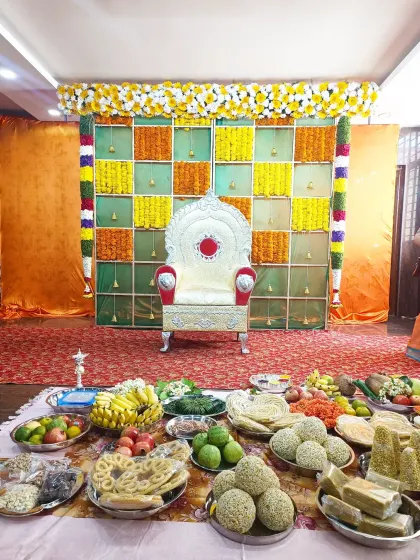 A colorful and traditional setup for a Shreemantha ceremony. The backdrop is a checkered pattern of marigold garlands, and the foreground is filled with fruits, sweets, and other traditional offerings.
