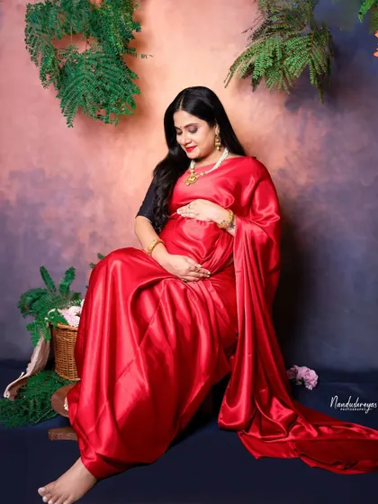 A serene portrait of the mother-to-be seated amidst green plants, her red saree a vibrant splash of color.