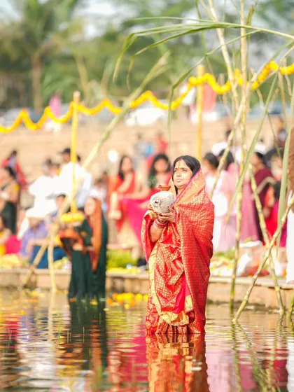 A woman in a red saree holds a coconut as part of her offering during Chhath Puja. The reflections in the water create a serene and beautiful scene.