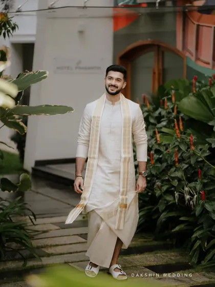 A smiling groom in a traditional South Indian outfit, walking through a garden.