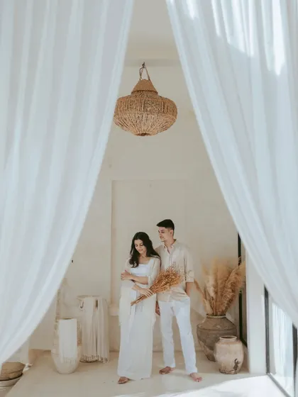 Framed by flowing white curtains, the couple stands together in a moment of quiet contemplation. The natural textures of the decor and soft lighting in this Bali villa create a warm and organic atmosphere.