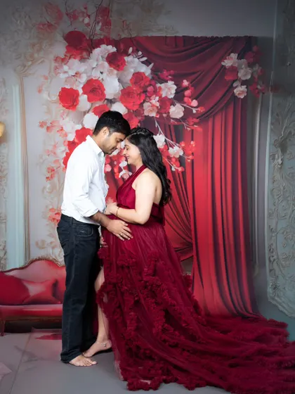 A romantic maternity portrait of a couple, forehead to forehead, surrounded by a dramatic red floral studio setup.