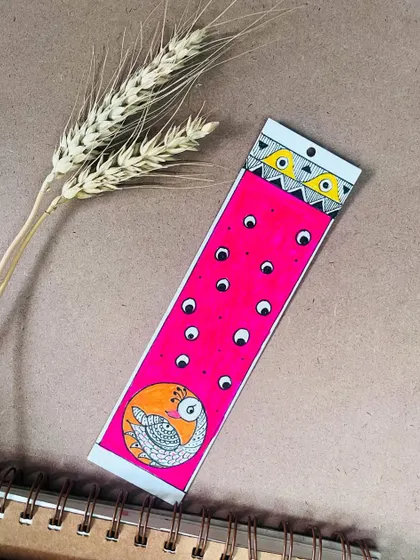 A bright pink bookmark featuring a Madhubani peacock design.