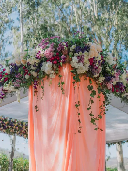 A close-up of the floral arrangement atop a mandap, showing the lush combination of flowers and cascading greenery against a soft peach drape.