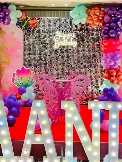 A silver shimmer wall provides a sparkling background for this fairy-themed birthday, enhanced by a "Happy Birthday" neon sign.