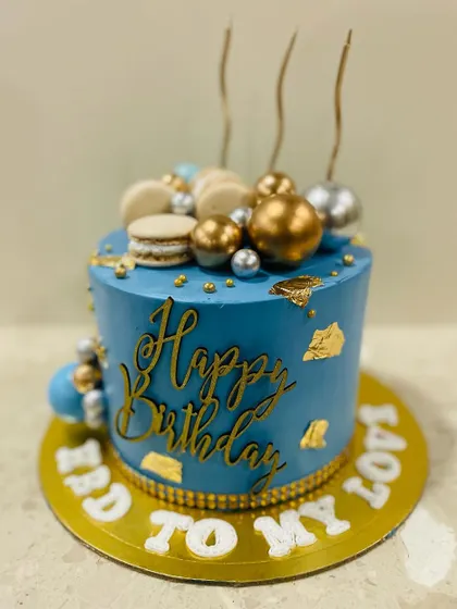 A sophisticated blue cake with gold calligraphy, topped with macarons and gold and silver spheres.