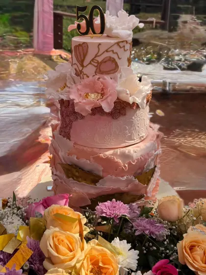 A three-tier cake designed for a 50th anniversary, featuring shades of pink, gold leaf, and delicate sugar flowers. The textured finish on the bottom tiers adds depth and elegance.