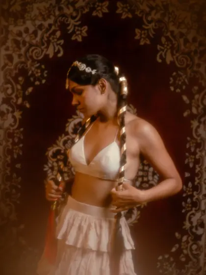 A portrait from the 'Lila' series, which has a vintage, bohemian feel. The styling includes a tiered skirt, a simple bralette, and hair wrapped with gold ribbon, set against a rich tapestry backdrop.