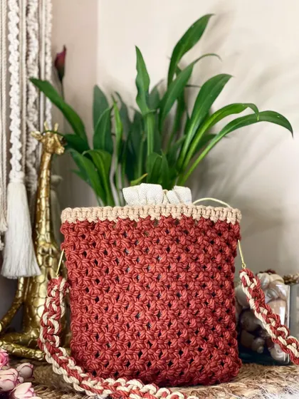 This rust-colored bag features a classic macrame net pattern and a secure drawstring top. The strap is woven with both rust and cream cords for a coordinated look.