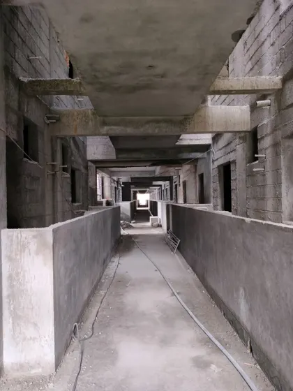 A long corridor inside a building during the raw construction phase. The exposed concrete ceiling, beams, and block walls show the building's structural shell before any finishing work has begun.
