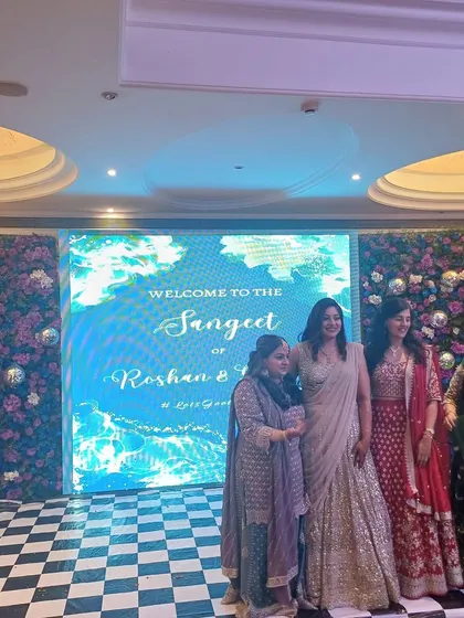Guests posing in front of a Sangeet stage with a large LED screen and floral walls.