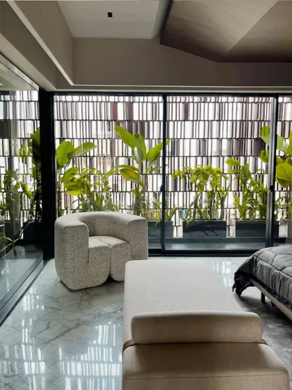 This bedroom corner is designed to embrace openness, with pristine white marble floors and expansive glass windows that connect to a balcony filled with lush greenery. The minimalist armchair and bench create an inviting retreat with an indoor-outdoor feel.