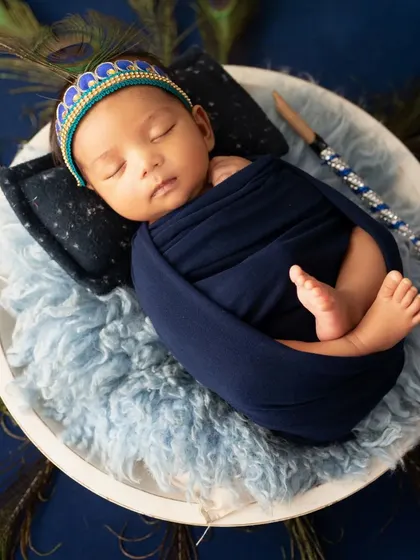 A sleeping newborn dressed as Krishna, wrapped in a blue swaddle and resting on a fluffy surface.