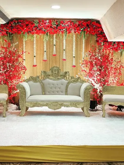 A bold and classic reception stage at the Crystal Hall in Acres Club. The combination of a rich red floral backdrop, traditional white and gold furniture, and elegant chandeliers creates a regal look.