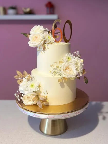 Simple, elegant, and timeless. This two-tiered 60th birthday cake in white and gold is adorned with fresh white roses and a touch of gold leaf, proving that less is often more.