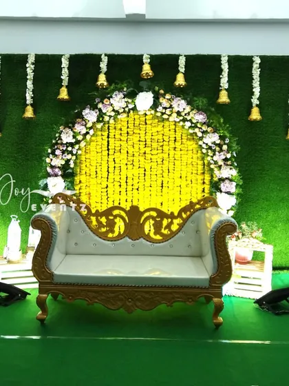 A beautiful stage setup for a Langa Voni function featuring a green turf backdrop and a floral arch. The design includes a yellow marigold curtain and hanging temple bells for a divine touch.