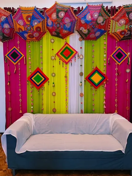 A festive Mehendi backdrop with colorful drapes in pink and lime green, adorned with traditional Rajasthani umbrellas.
