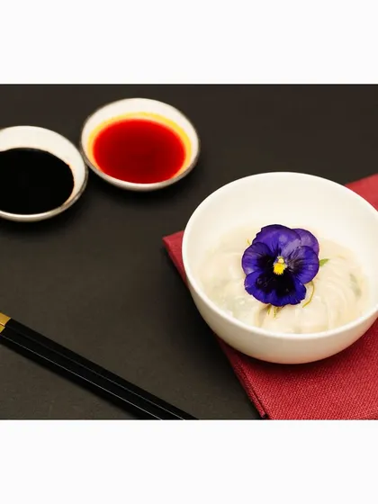 A delicate bowl of dumplings garnished with an edible purple flower, served with soy and chili sauces. This shot focuses on the fine details and beautiful plating of gourmet Asian cuisine.