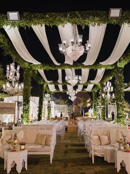 A symphony of florals and light. This elegant outdoor wedding seating is arranged under a canopy of green garlands and white drapes, with crystal chandeliers adding a touch of romance.