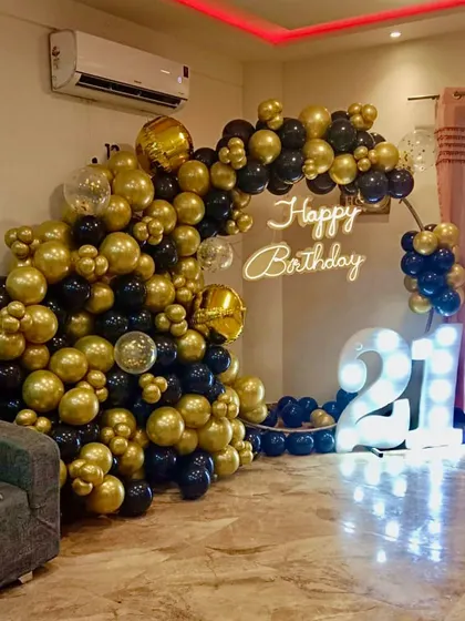 Celebrating a 21st birthday in style. This setup features a dramatic black and gold organic balloon arch, a custom 'Happy Birthday' neon sign, and large, illuminated marquee numbers to mark the special occasion in any home.