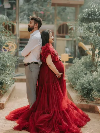A creative and intimate pose for a couple's maternity shoot. The rich red of the ball gown stands out beautifully against the green garden background.