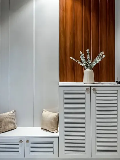 A minimalist foyer featuring a white shoe cabinet with louvered doors for ventilation and a matching bench. The design is complemented by a wood-paneled accent wall, adding warmth and texture to the space.