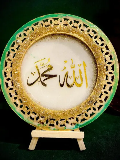 A circular tabletop plaque with 'Allah & Mohammed' in gold calligraphy. The piece has a decorative green and gold filigree border, giving it a traditional yet modern look.