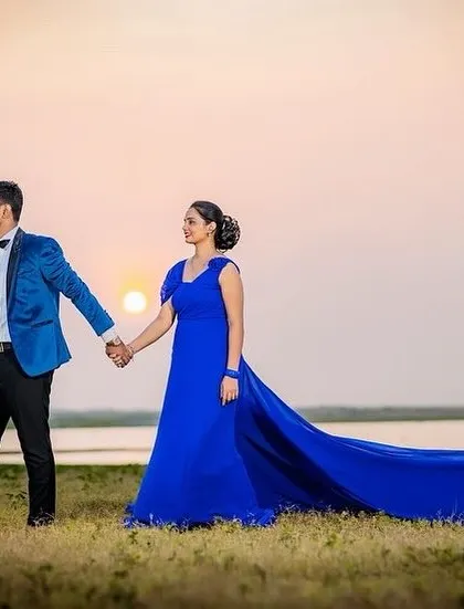 A sunset pre-wedding shoot by the water. The groom-to-be is wearing a stylish blue velvet tuxedo jacket that perfectly complements her royal blue trail gown.