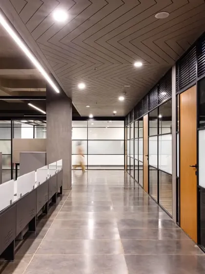 A view down the corridor of the workstation area, showing the glass-walled private cabins and the detailed ceiling with a herringbone pattern. The design optimizes both individual productivity and team interaction.