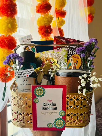 A festive Rakhi hamper in a golden basket, held against a backdrop of marigold garlands. It includes The Body Shop products and snacks.