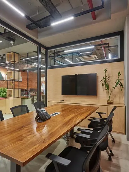 A meeting room with an exposed brick wall and a large wooden table, viewed from the outside. The glass partitions allow for visual continuity with the adjacent reception area.