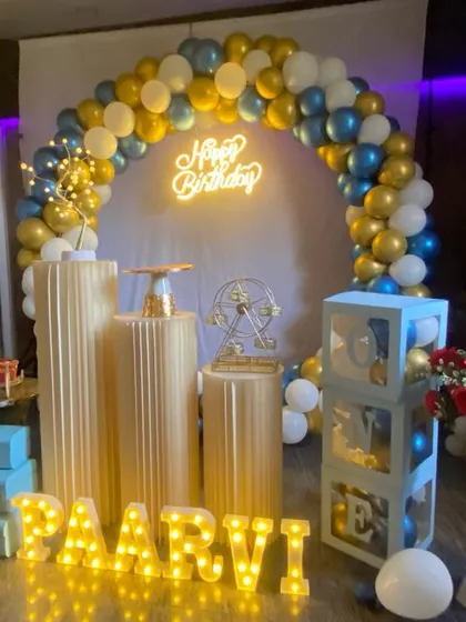 A birthday setup with a gold, blue, and white balloon arch. The decor features gold plinths, 'LOVE' blocks, and marquee letters for the name.