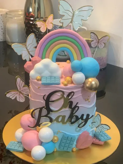 This "Oh Baby" cake is perfect for a baby shower or gender reveal party. The design features a beautiful pastel rainbow, fluffy clouds, and a mix of pink, blue, and gold chocolate spheres.