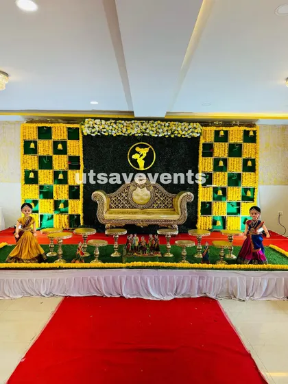 A full view of the baby shower stage with a grass wall and marigold backdrop. The red carpet runner leads up to the ornate golden sofa, making the setup look grand.