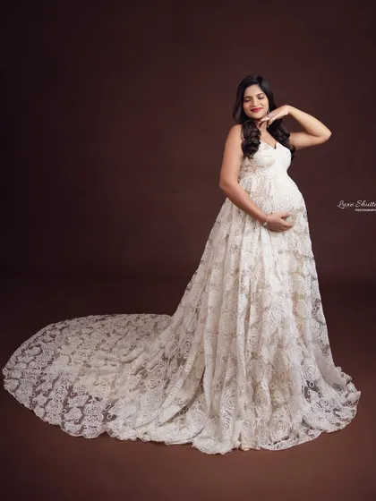 The intricate detail of this white lace gown is simply breathtaking. It adds a layer of texture and vintage-inspired elegance to the maternity portrait, set against a classic studio backdrop.