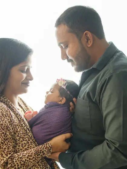A beautiful portrait of new parents admiring their sleeping baby, wrapped in a deep purple swaddle. The lighting highlights their features and the baby's peacefulness.