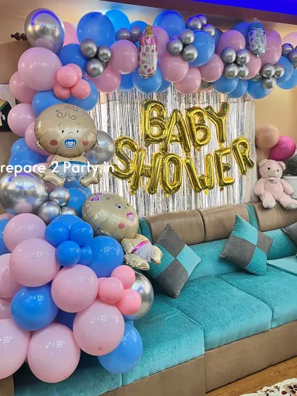 A classic at-home baby shower decoration. The setup features a pink and blue balloon arch over a couch, a silver shimmer curtain, and large gold foil letters spelling "BABY SHOWER".
