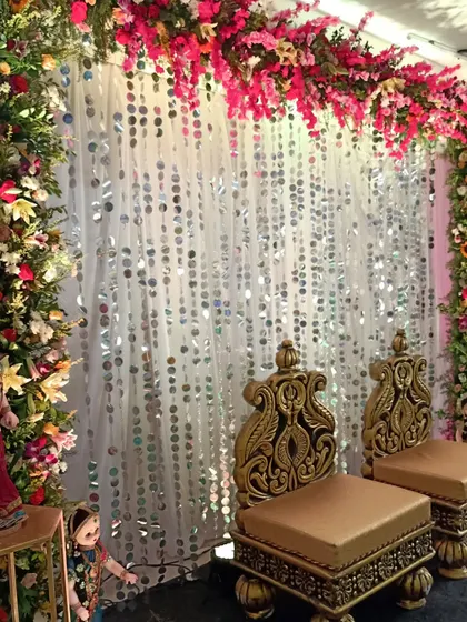 This Haldi stage combines tradition with a touch of glamour. The backdrop features a shimmering sequin curtain framed by colorful floral arrangements, with ornate golden chairs for the couple.