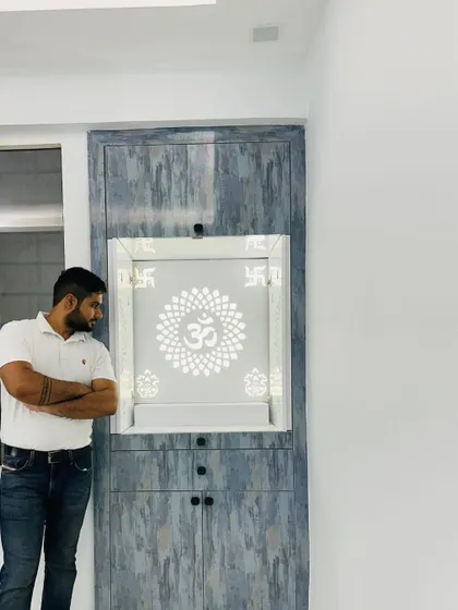 Here I am next to a modern mandir unit. The design features a grey wood-finish laminate and a backlit Corian panel with a beautiful 'Om' design, creating a contemporary yet spiritual space.